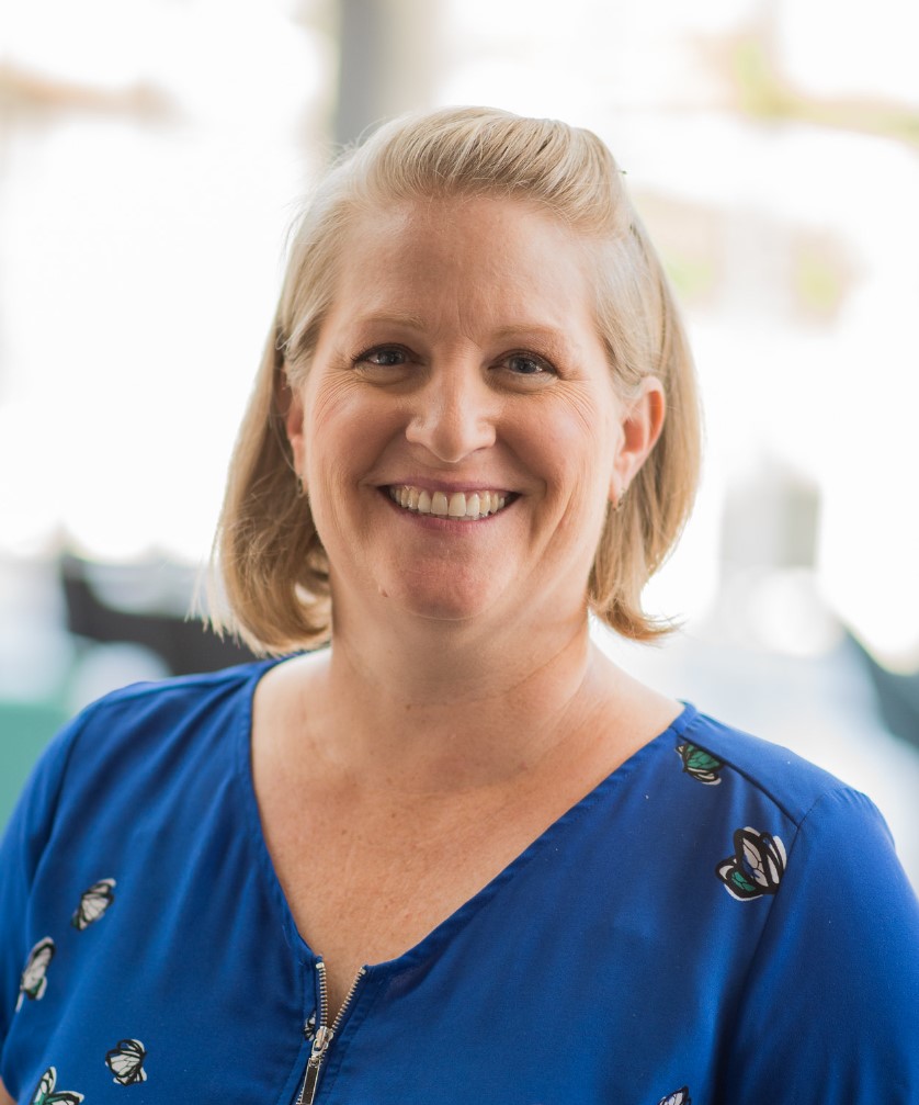 Nicole Mart has short blond hair and is wearing a blue blouse with white and green flowers. She is smiling in front of a bright blurry background.