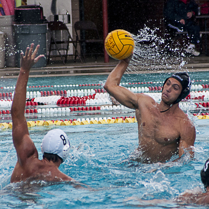 A male Palomar water polo player tries to throw a ball from as an opponent tries to block him with his left arm.