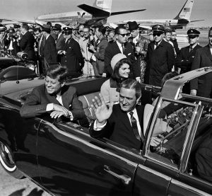 President John F. Kennedy and First Lady Jacqueline O. Kennedy sit in the back seat of a convertible car . Texas Gov. John Connally sits in the passenger seat, waving with his right hand. Several Secret Servicemen and reporters and two commercial airplanes are in the background. Black and white photo.