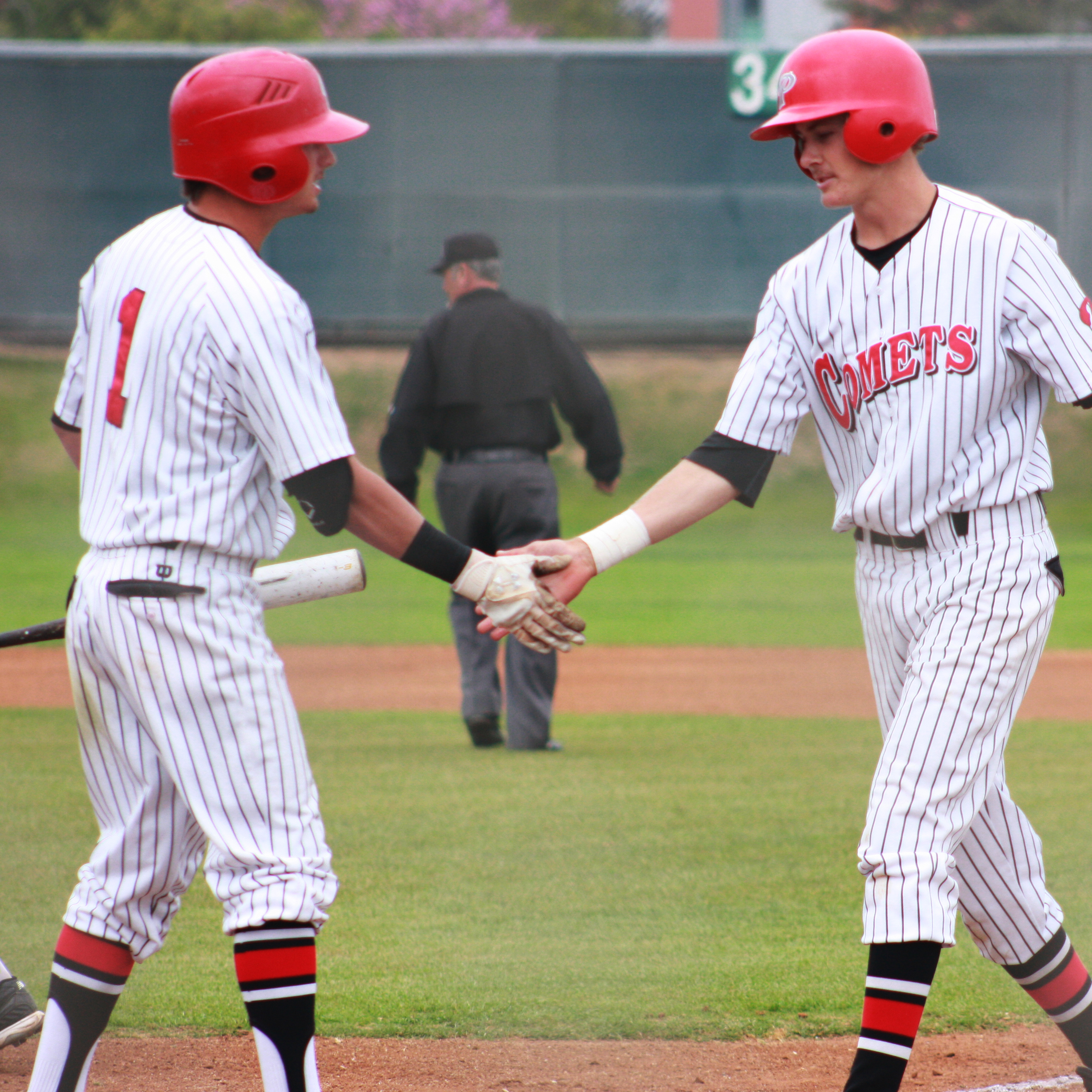 Two Palomar baseball players shake hands on the one on the right walks toward the player on the left. An umpire walks away from them in the background.