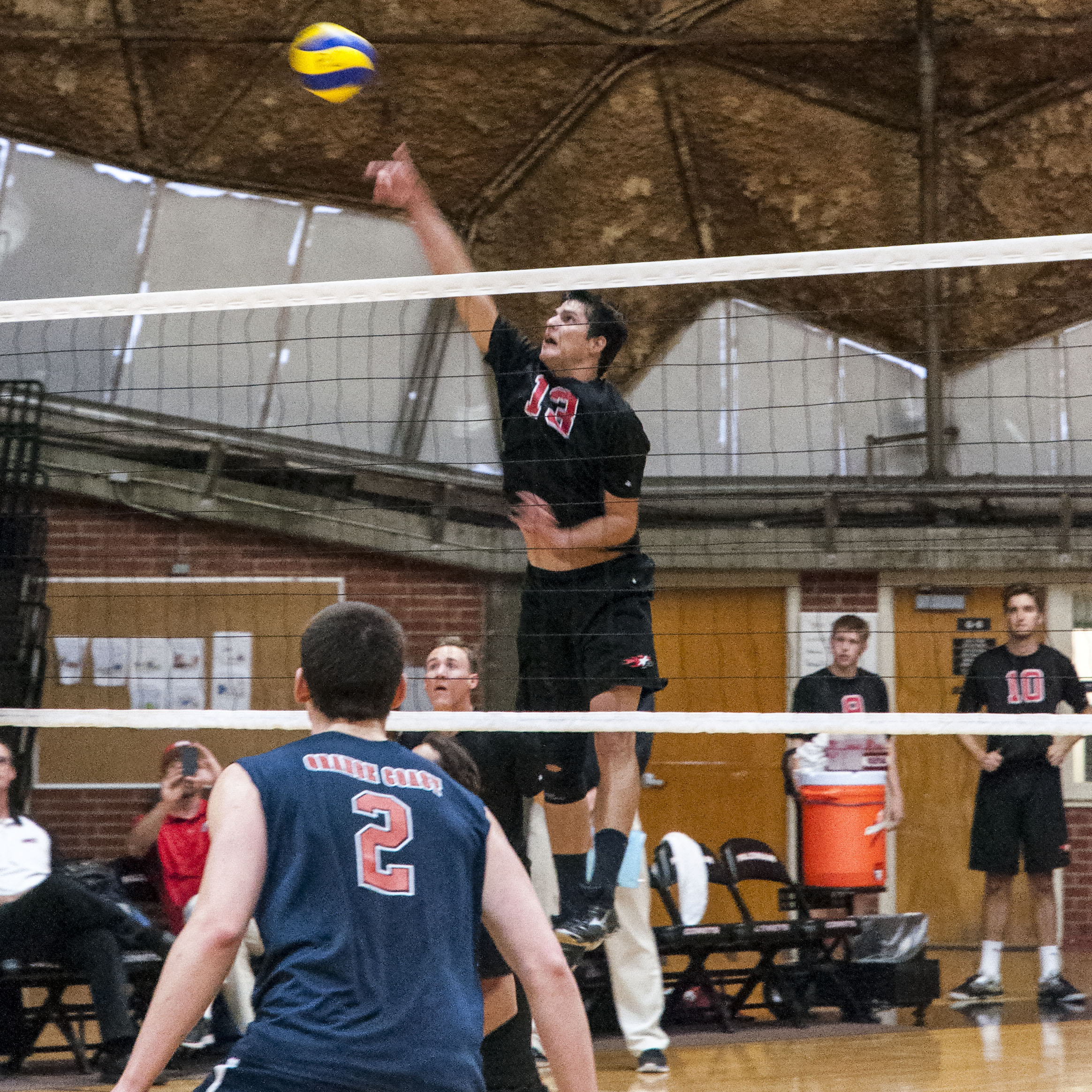 A male Palomar volleyball player jumps and hits the ball over the net as an opponent in the foreground watches.