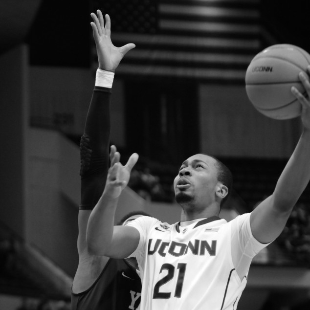 A male basketball player does a lay-up with his left hand holding a basketball. An opponent tries to block him with his right hand over his head. Black and white photo.