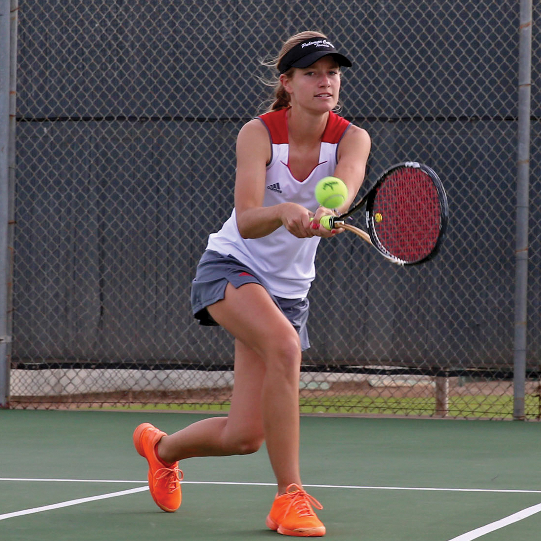 Palomar tennis player Remy Littrel swings a tennis racket with both hands at a tennis ball in a tennis court. She wears a white sleeveless jersey with red on the upper shoulders, gray shorts, a black visor, and orange shoes.
