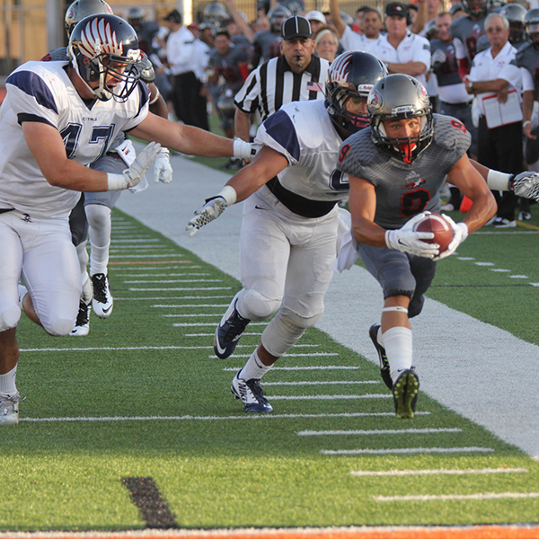 A Palomar football player runs near the sidelines as two opposing players chase him from behind and to his right.