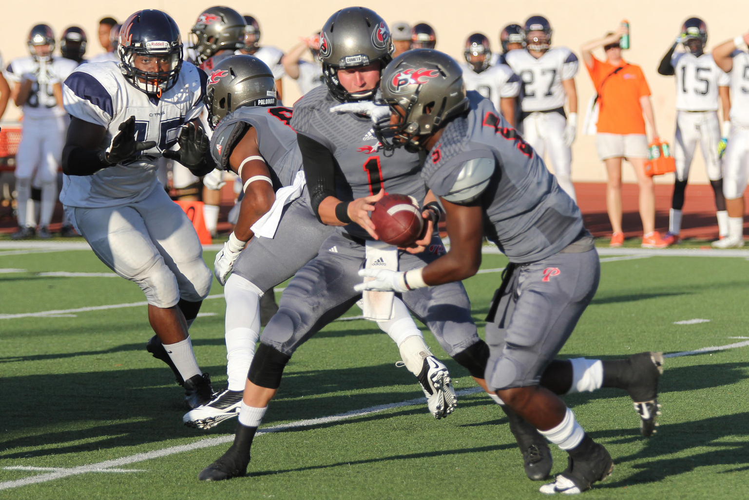 A Palomar football player hands a team player a football as another team player tries to block an opposing player on the left.