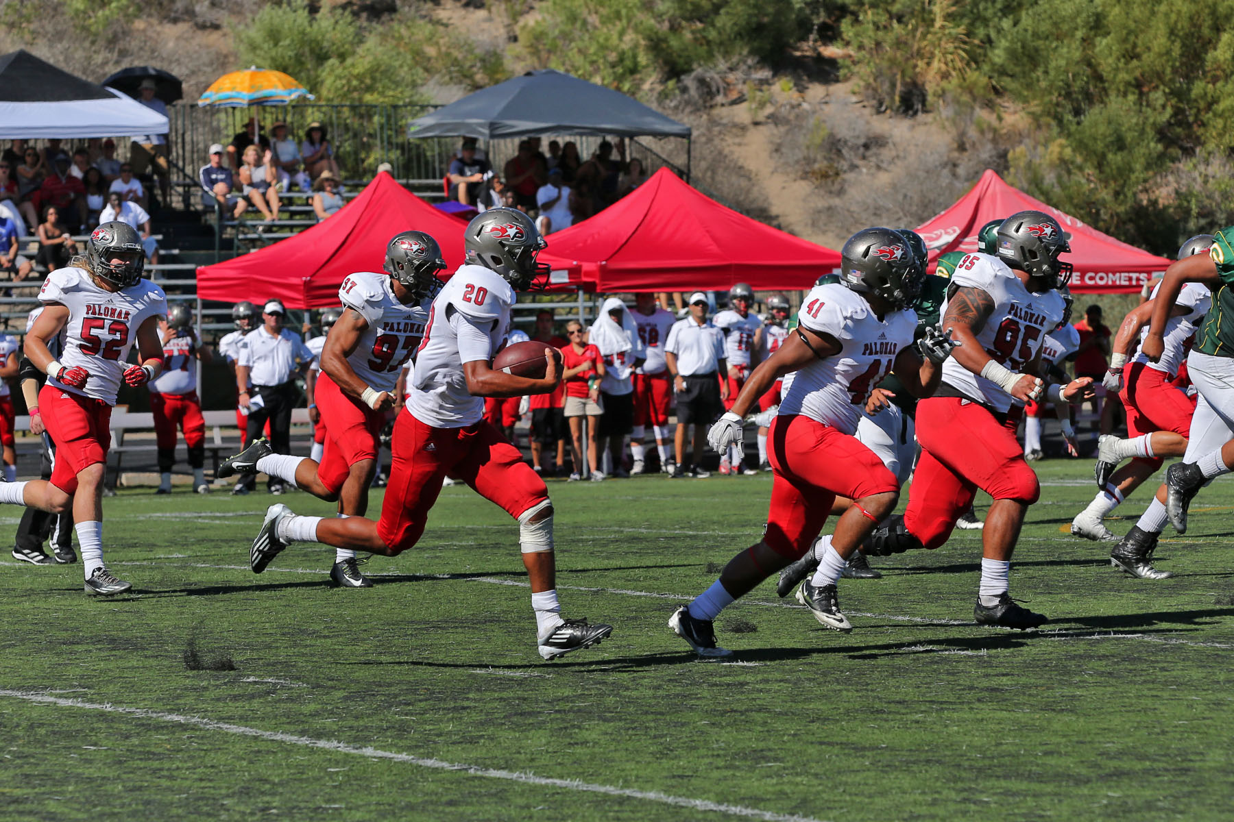 A group of Palomar football players run across a field with the middle player carrying a football in his arms. A crowd of people watch from the bleachers and tents in the background.