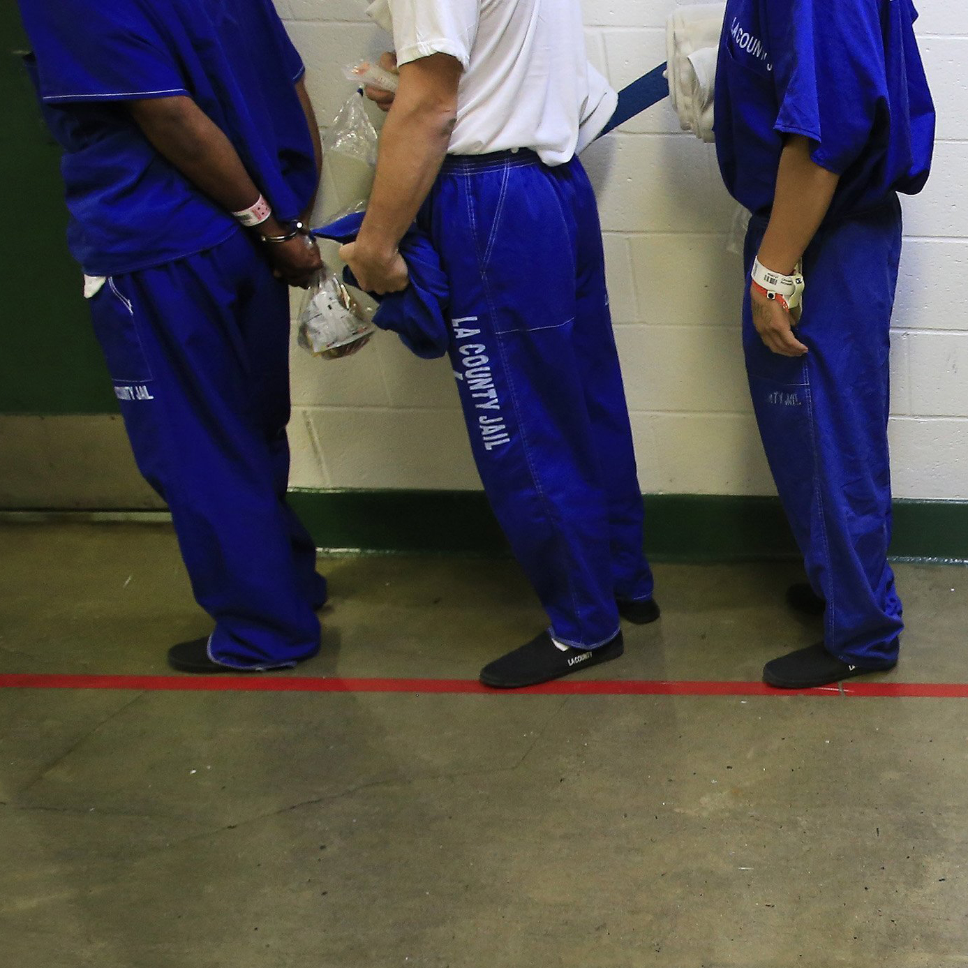 Three male inmates stand in a queue, wearing blue pants and blue shirts, except for the middle person who is wearing a white undershirt. They are seen only from the lower torso to their feet.
