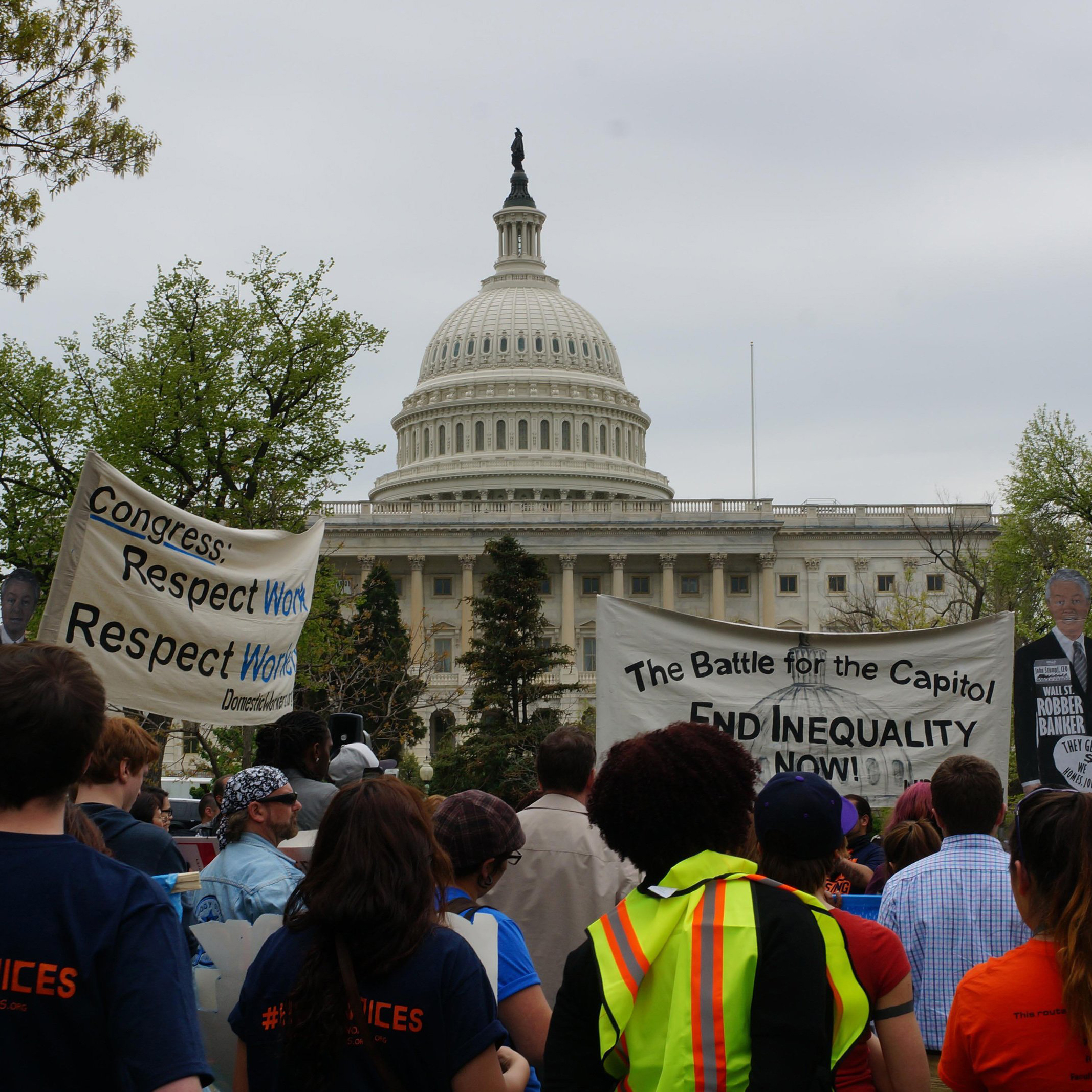 A group of protesters stand outside of the U.S. Capitol buidling on a cloudy day. A sign on the left reads, "Congress: Respect Work Respect Work." A sign on the right reads, "The Battle for the Capitol. End Inequality Now!"