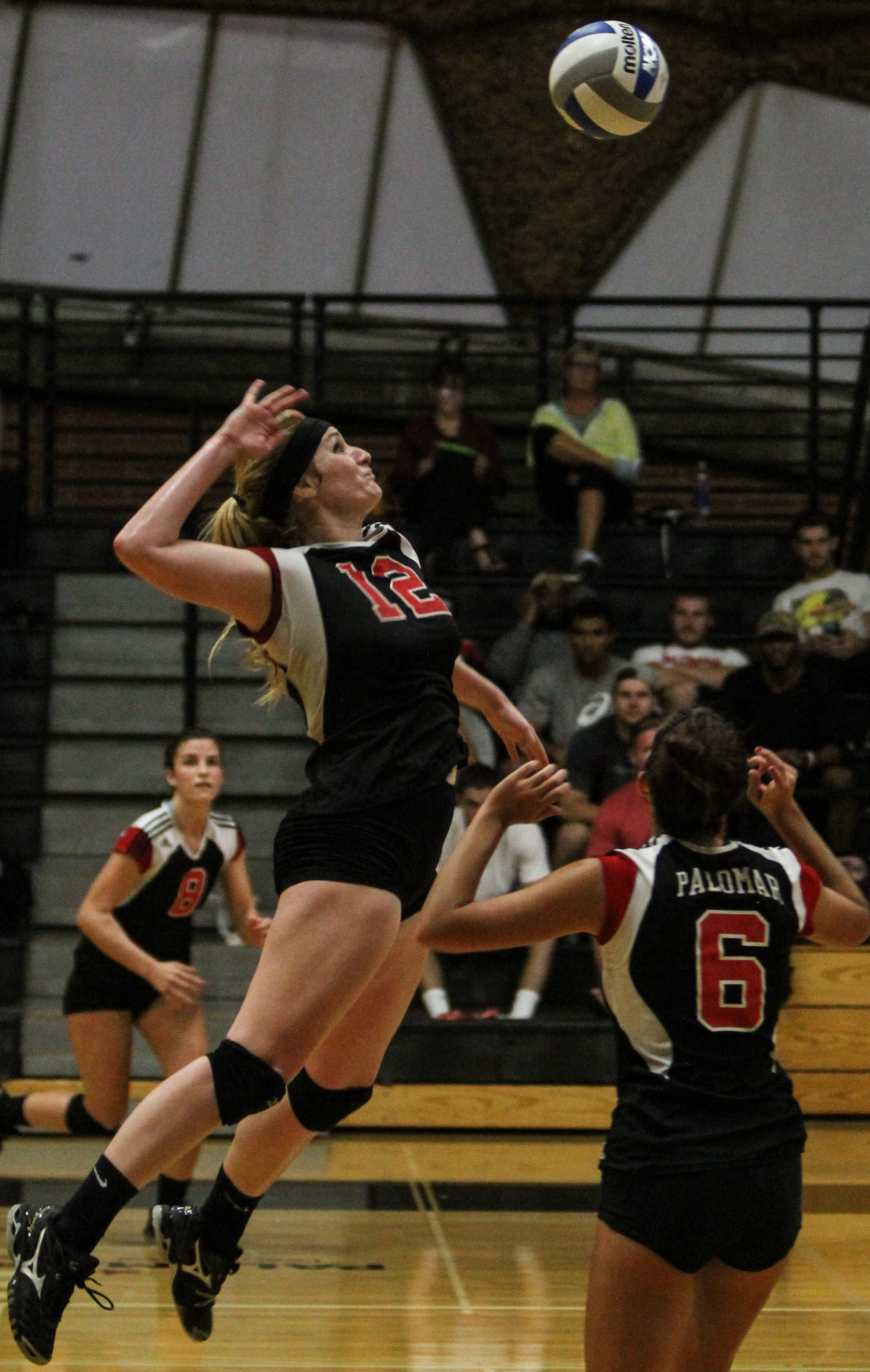 A female Palomar volleyball player leaps up to serve the ball with her right hand. A teammate stands near her on the right.