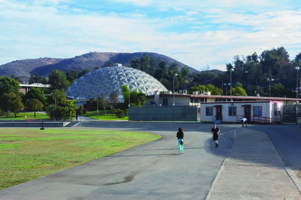 A running track with a grassy football field on the left and The Dome in the background. A bungalow in the right in the middleground. Two people jog on the track while a man sits on a lawn chair near the bungalow.