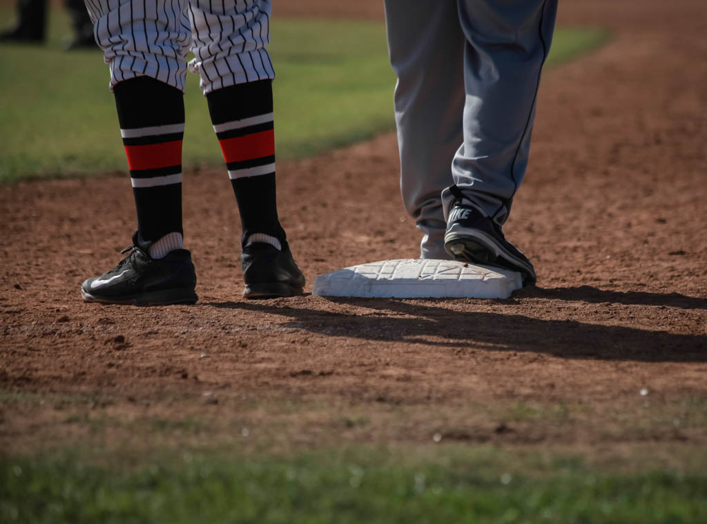 Two pairs of lower legs and feet in baseball shoes stand near a base on a field. The pair on the left wears long black socks with a thick red stripe and a black and white stripe above and below it. The pair on the right wears long gray sport pants.
