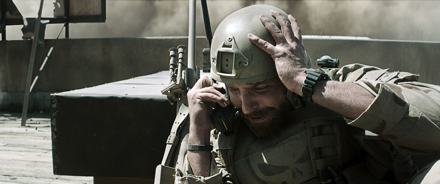 Bradley Cooper as Chris Kylethe movie "American Sniper." He wears dark green military gear and helmoet, his left hand on his head while holding a walkie-talkie in his left hand.