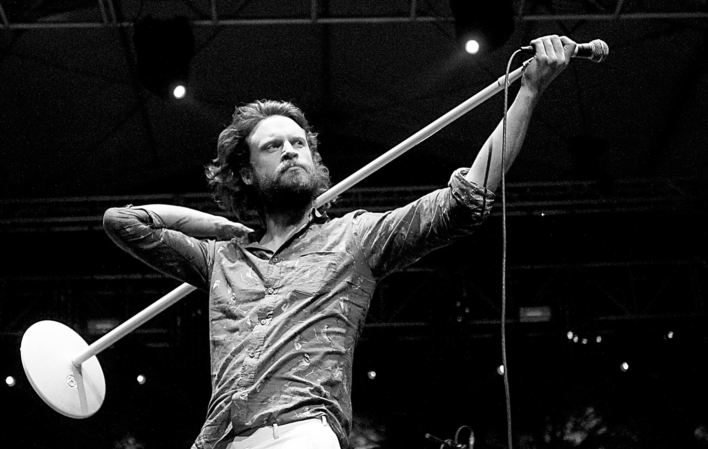Father John Misty holds a microphone and its stands with both hands.