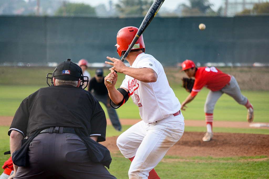 A male Palomar baseball player prepares to swing a bat as the pitcher throws a baseball toward him. An umpire stands and leans forward on the lower left, his bottom facing the viewer.