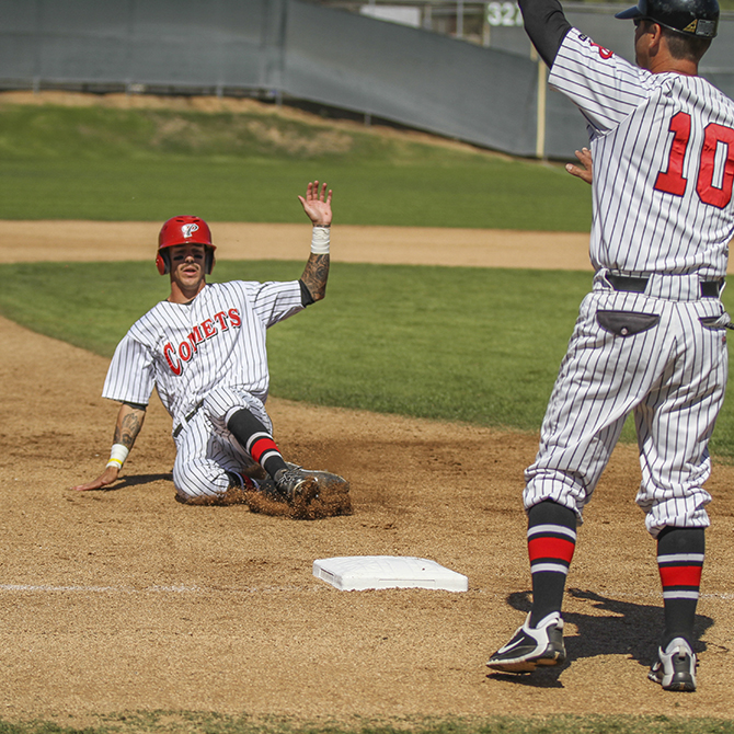 A male Palomar baseball player slides to base on his bottom with his left hand raised and left elbow bent. A fellow player stands in front of him by the base, raising his left hand.