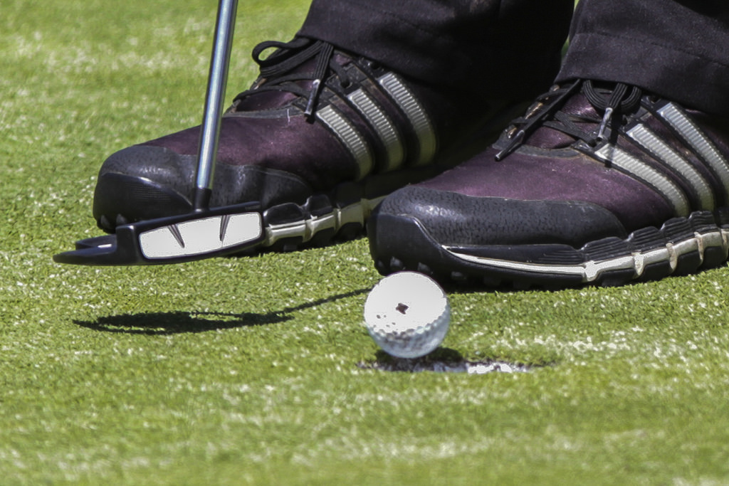 A pair of feet in purple and black shoes with white lines stand close to a golf golf that is on the edge of a hole. A golf club head hovers above the turf in front of the shoes.