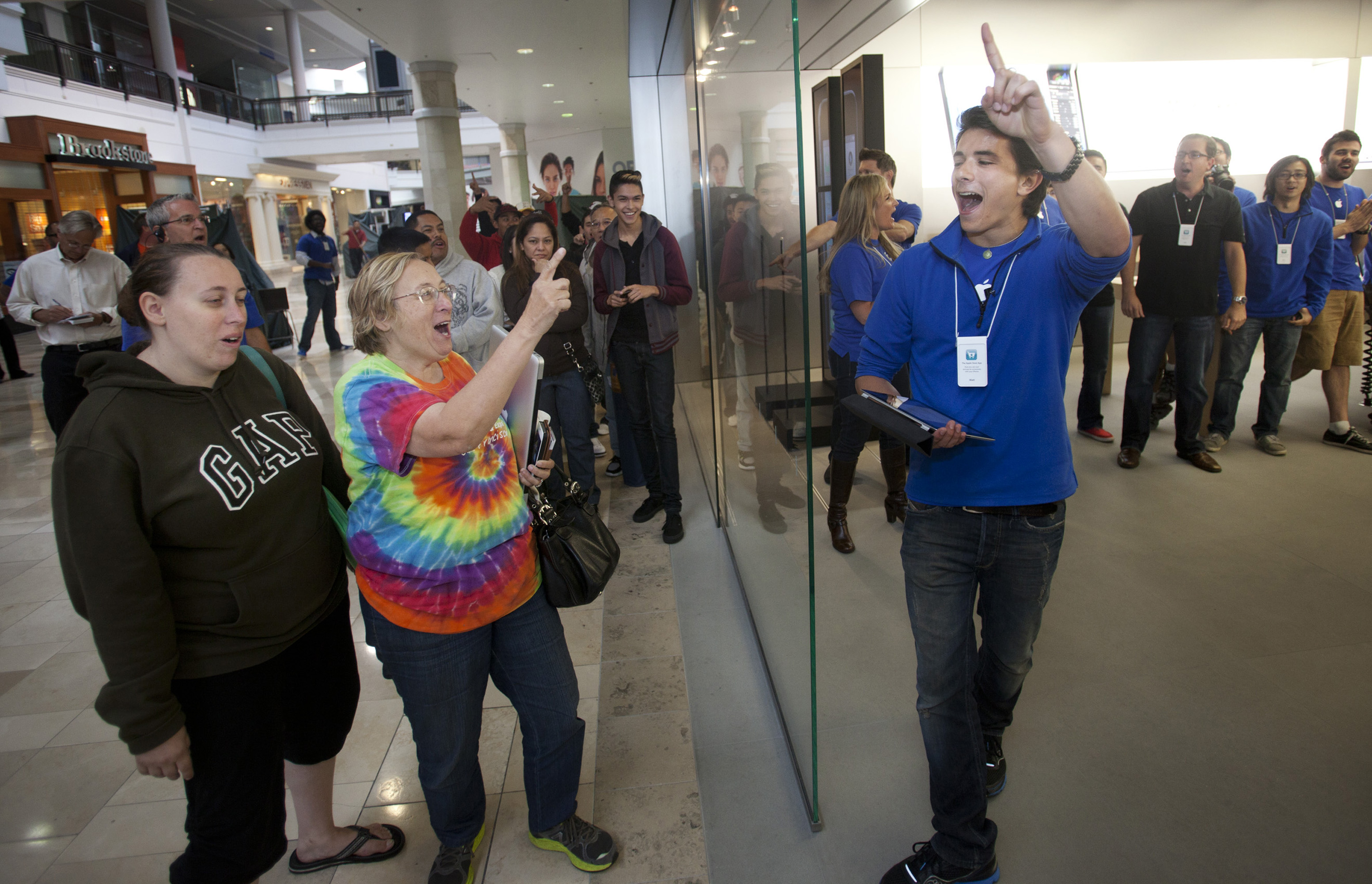 A group of people wait outside of an Apple store on the left while a group of Apple store employee stand on the right. One of the male employees stand next to the glass door holding a clipboard and his left finger up as he talks (or shouts) with a woman outside on the left.