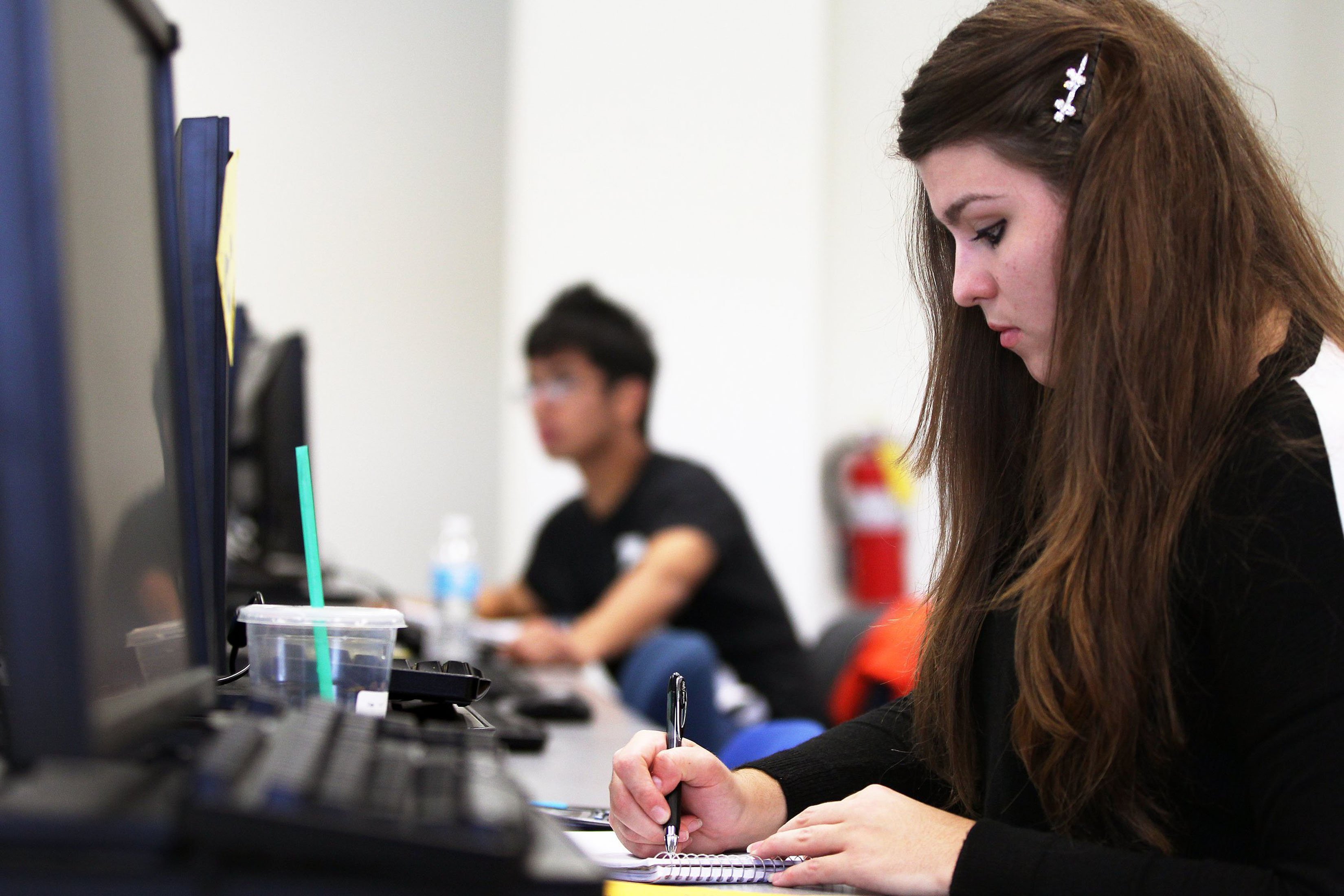 A young, female student writes in her notebook in front of a computer monitor and keyboard with a Starbucks drink near her. Another male student sits in the background (blurred).