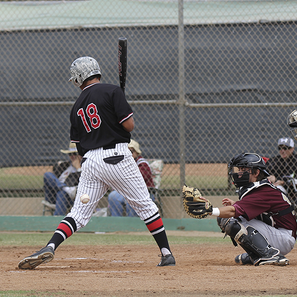 A male Palomar baseball player shifts his weight to his right leg as a baseball nearly hits his left leg. A catcher squats behind him with his left mitted hand extended to catch the ball.