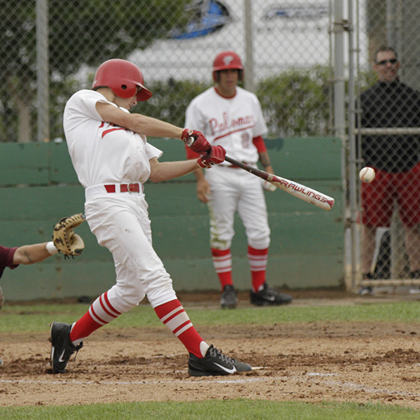 A male Palomar baseball player swing a bat and hits a baseball to the right. A catcher mitted hand is at the lower left by the player's butt. Another team player and a man in a black sweater and red shorts stand in the background, watching the game.