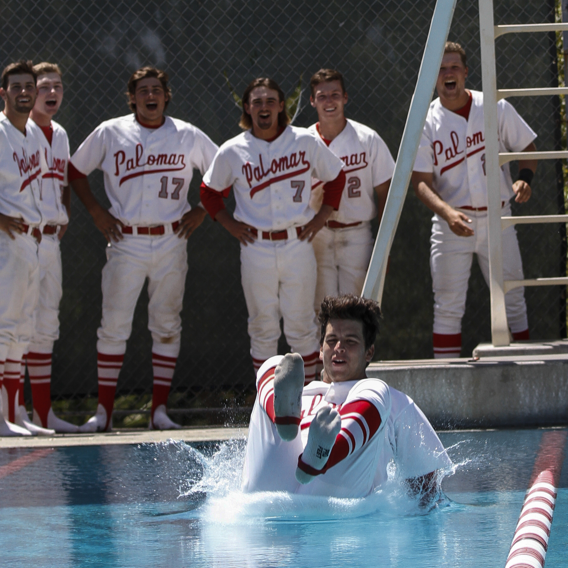 A male Palomar baseball player jumps into a pool butt-first with his feet in socks facing the viewer. Six team players stand in the background and laugh.