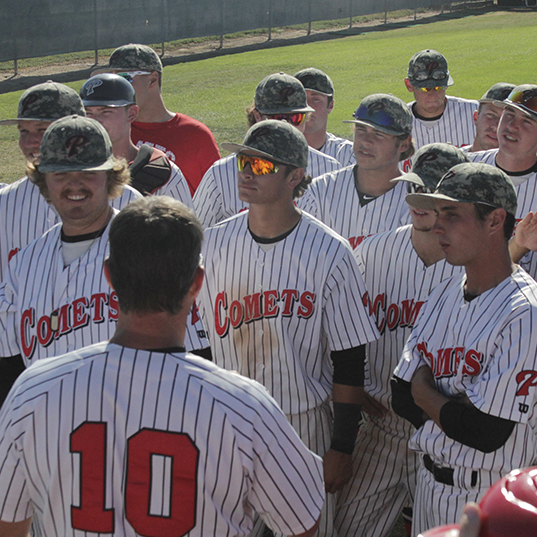 A group of male baseball players stand and gather around their coach on a sunny day in the field. They all wear white jerseys with think vertical striples with the word "COMETS" in the front. Only one guy in the background wears a red T-shirt.