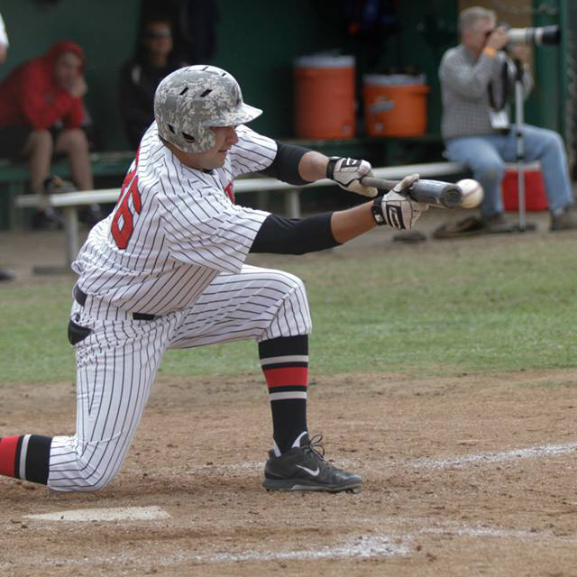 A Palomar baseball player kneels on his right knee and bunts a baseball which is almost touching the bat. A photographer is in the background on the right and two people watch the game in the dugout.