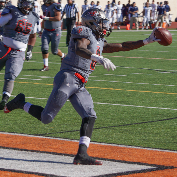 A Palomar football player runs while holding a football in his left hand. Several team players run alongside near his left.