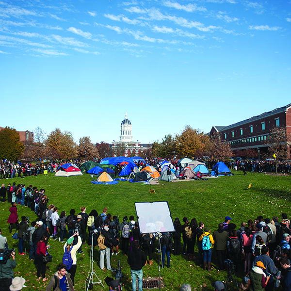 A crowd student protesters surround a group of tents pitched in the middle of a grassy plaza in a university.