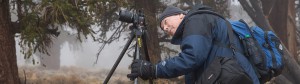 Palomar photography student Terry Ogden bends forward and peers through a camera on a tripod. He wears a blue and black coat and a fade blue beanie.