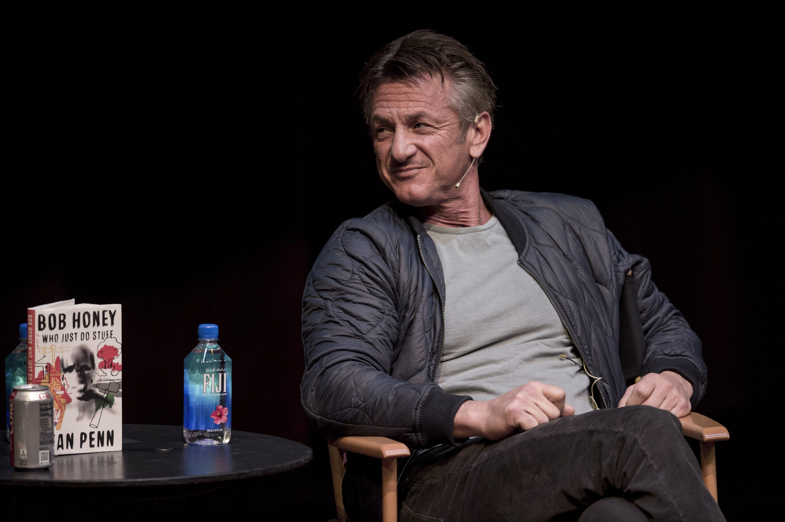 Sean Penn sits cross-legged and looks to his right, wearing an earpiece, a light navy-blue jacket, and a grey shirt. A bottle of Fiji water and a book titled, "Bob Honey Who Just Do Stuff," stand on a table to his right.