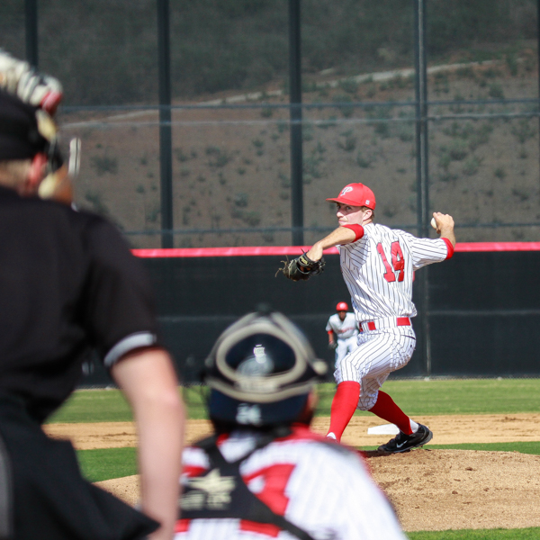 A male Palomar baseball player cocks his right arm back to pitch a baseball as a catcher and an umpire look at him in the foreground (blurry).