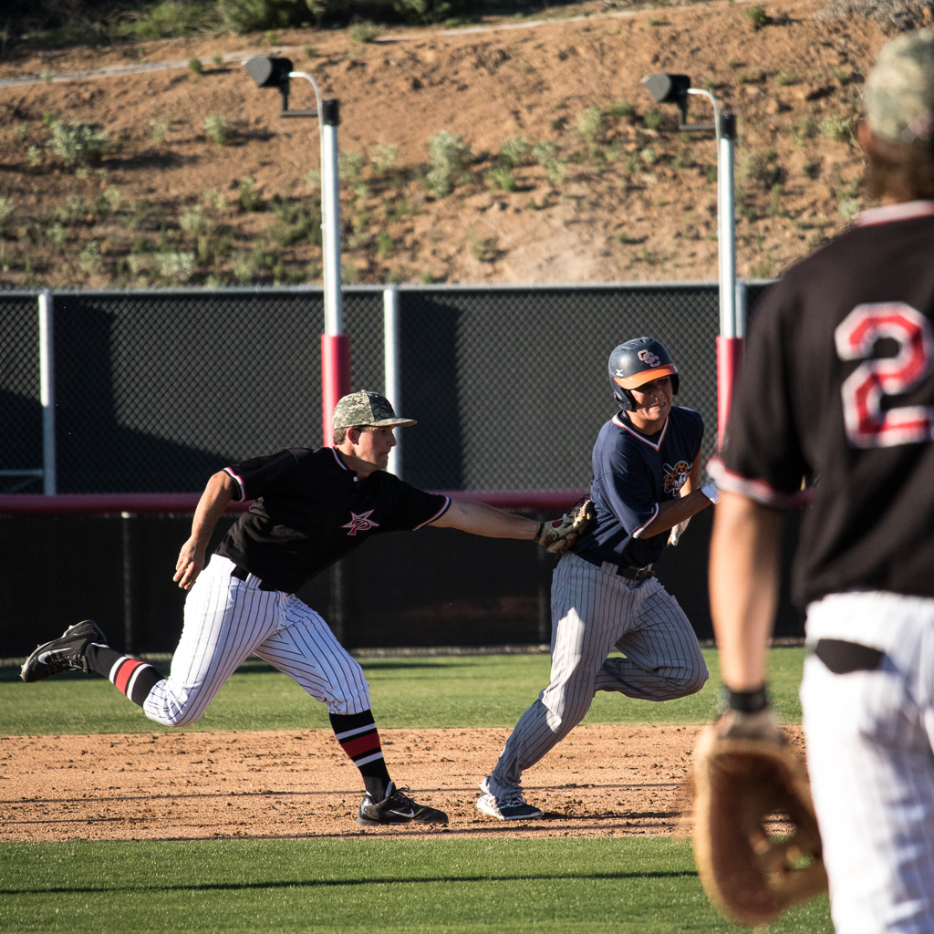 A male Palomar baseball player tags an opposing player as he runs after him. A teammate stands in the foreground (blurred), watching them.