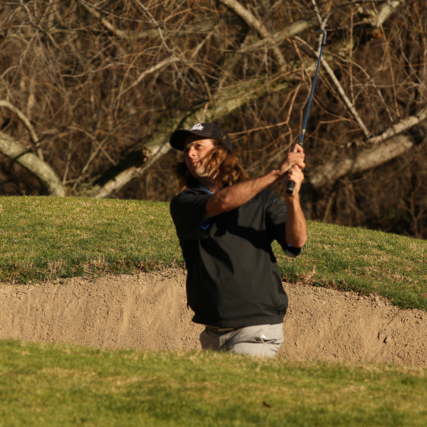 Palomar Golf player Kelly Cannon swings a golf club to knock a golf ball off the sand. He wears a black cap, a black shirt, and gray pants.