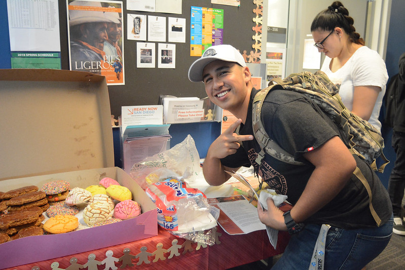 A male Palomar student leans forward and tilts to his right, giving the reverse peace sign near a big box of Mexican sweetbread. A female student stands behind him.