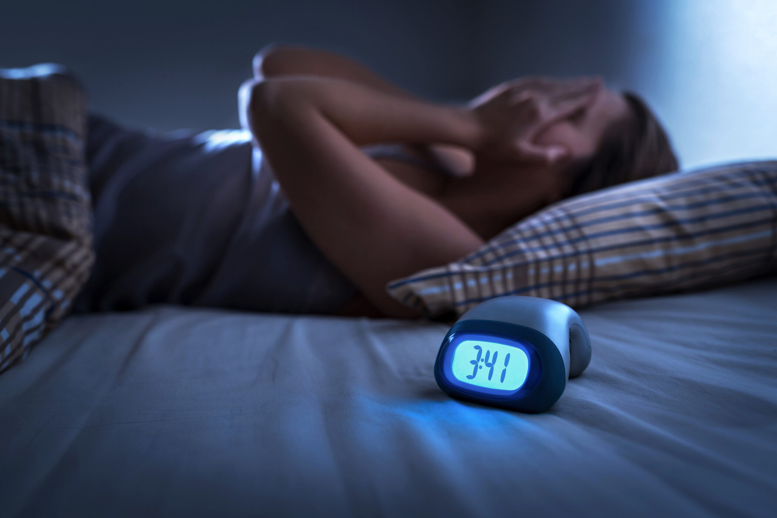 A blue digital clock displays the time 3:41 on a bed with a blurry image of a woman laying supine with her hands to her face.