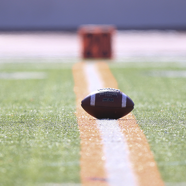 A football lies on a field on top of a white and orange line. An orange box (blurry) sits on the ground in the background.