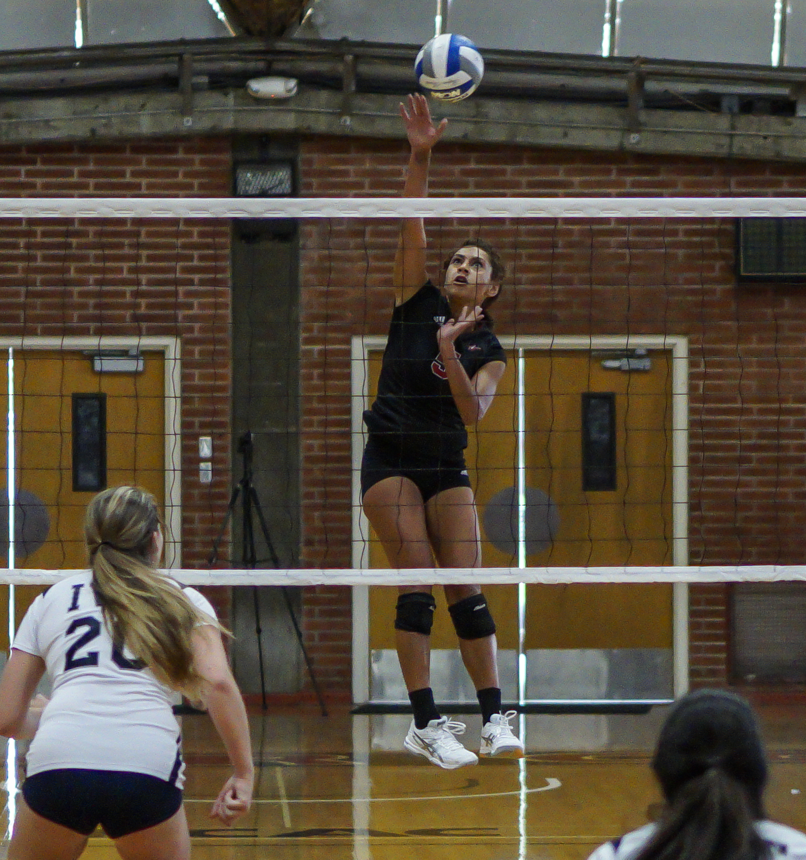 A female Palomar volleyball player jumps up and spikes the ball over a net with her right hand. Two opponents stand in the foreground.