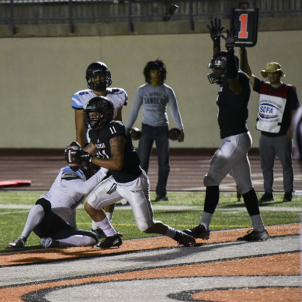 A Palomar football player runs with a football in both hands while an opposing player tries to drag him down in a sitting position. A team player walks behind them with his hands above his head. Several people stand on the sidelines in the background close to them.