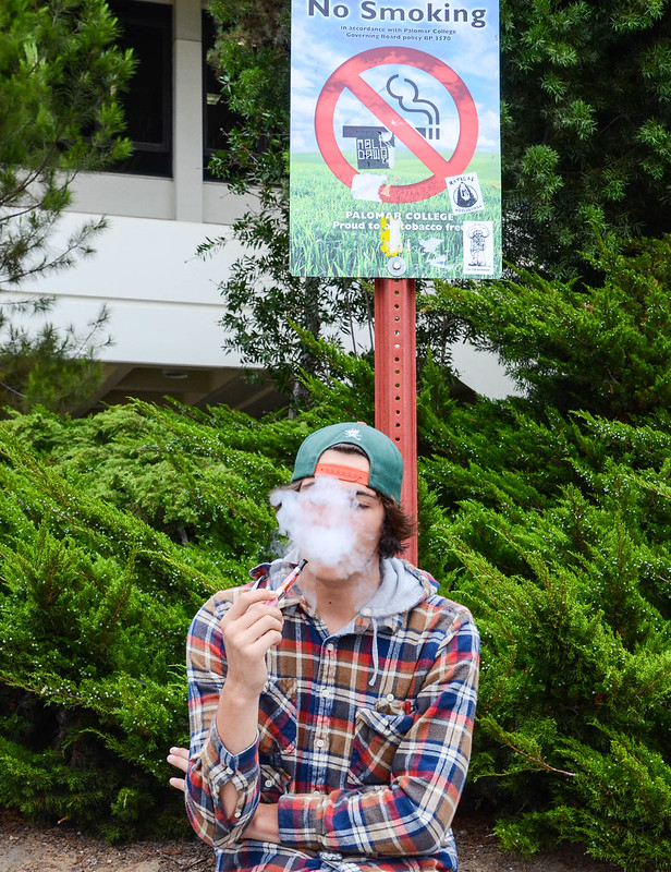 A male Palomar students sits and vapes beneath a "No Smoking" sign.