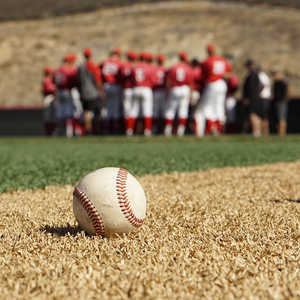 A baseball lies on yellow turf with a team of Palomar baseball players and coaches standing in the background (blurry).