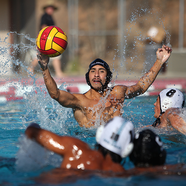 A male Palomar water polo player throws a ball with his right hand as several players swim in front of him in the foreground.