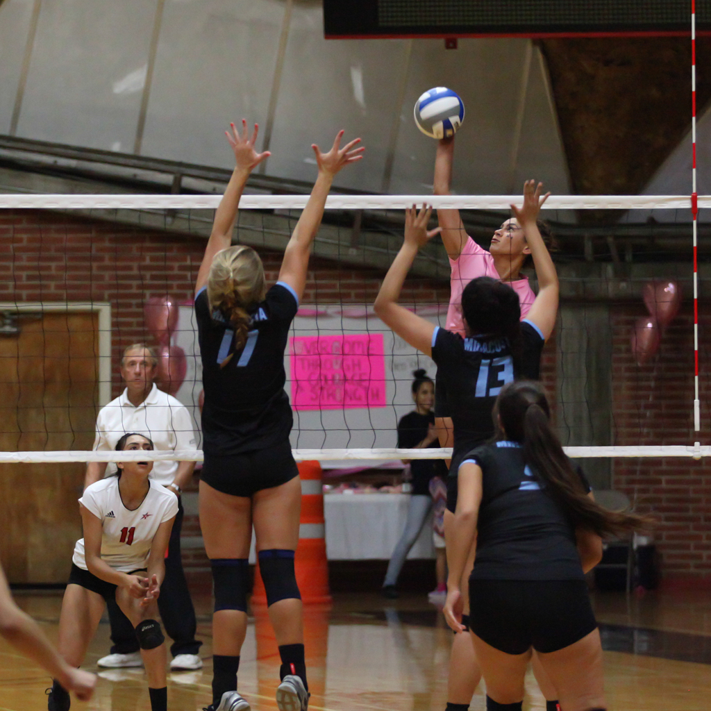 A female Palomar volleyball player spikes a ball as two opponents try to block it.