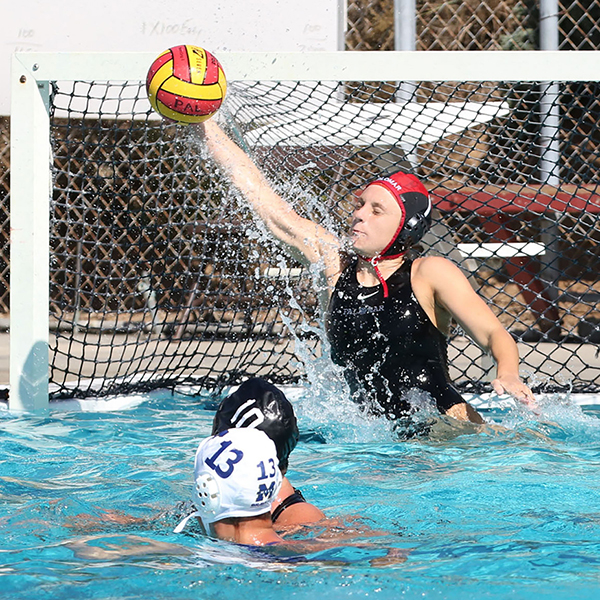 A female Palomar water polo player tries to block a ball from going into the goal with her right hand. Two players are swimming in front of her.