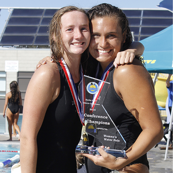 Two female Palomar water polo players hug and pose for the camera. One of them holds a 2016 Women's Water Polo PCAC Conference Champions trophy.