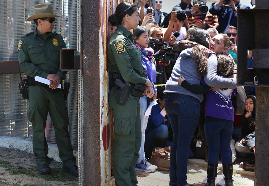 Two female border patrol agents in dark green uniforms stand at a gate as a crowd of people watch a mother and a young girl embrace on the right.