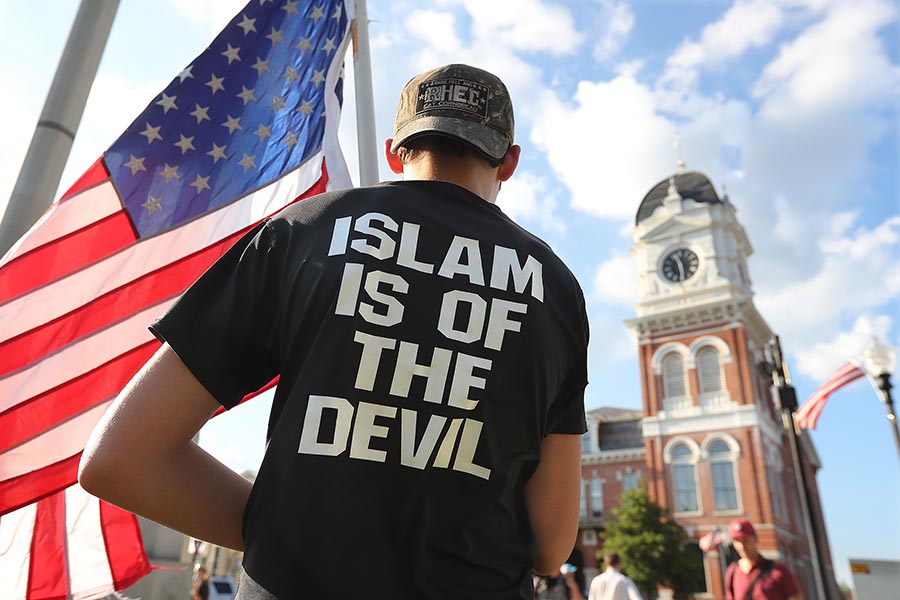 A man wears a black T-shirt that has the text on the back, "ISLAM IS OF THE DEVIL." He holds an American flag. A clocktower is on the right.