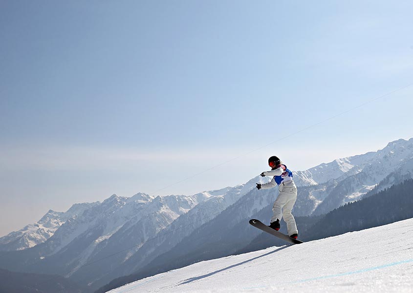 A snowboarder crusies downhill on a snowy mountain with her arms extended in front of her. A snowy mountain range is in the background.