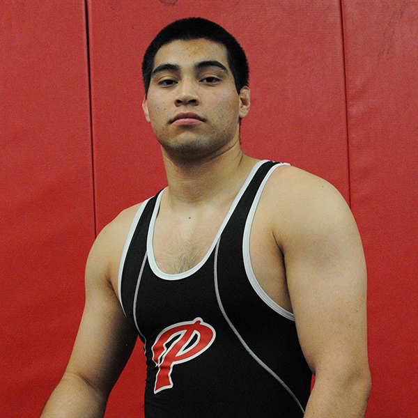 Palomar College wrestler Alex Gomez kneels on his left knee, wearing a black jersey, shorts, and wrestling shoes. Palomar's letter "P" logo is in the middle of the jersey.