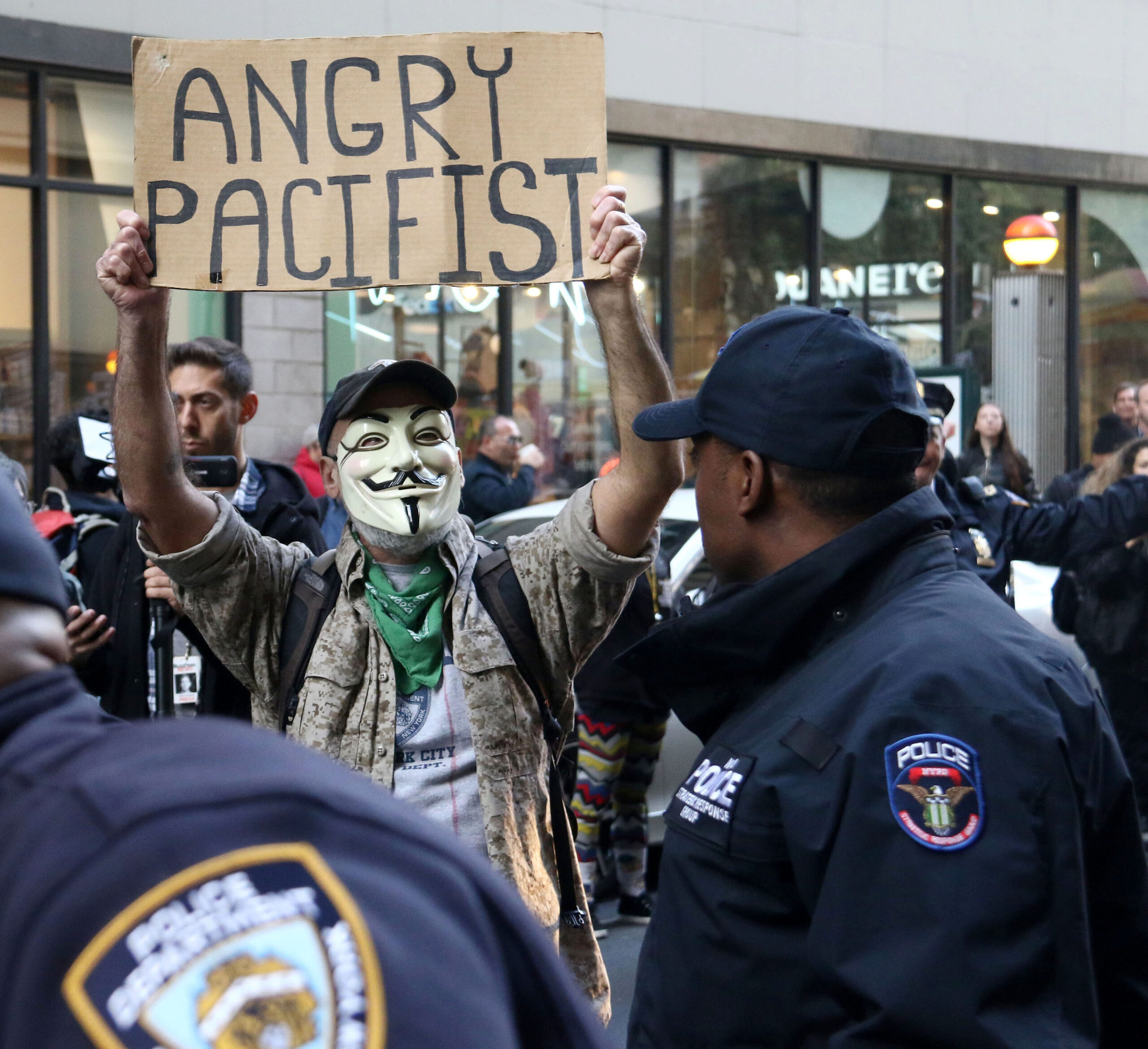 A protestor wearing a Guy Fawkes mask holds a cardboard sign that says, "Angry Pacifist." A police officer looks at him at the front.